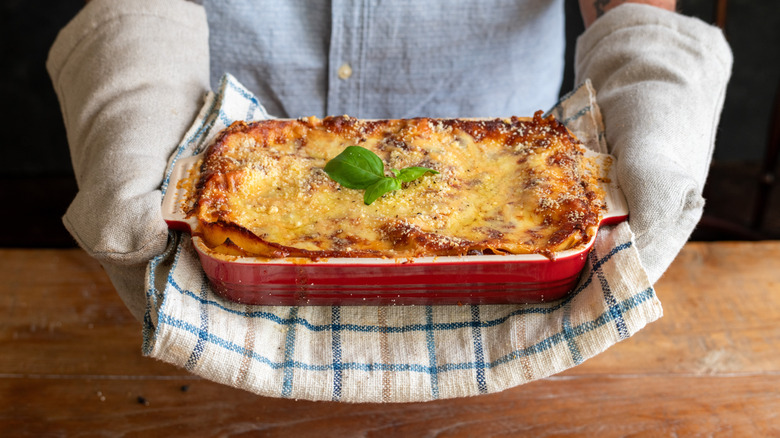 A person wearing oven mitts holding a red casserole dish featuring a cheese topped casserole with fresh basil leaves on it