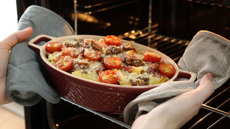 person taking out a meat, potato, tomato casserole from the oven using oven mitts