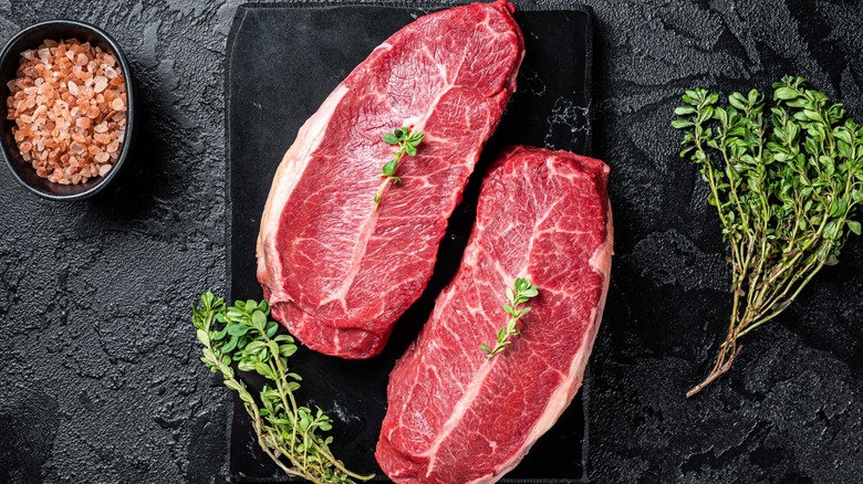 Raw charcoal or top blade steak on a black stone cutting board with fresh herbs and pink salt.