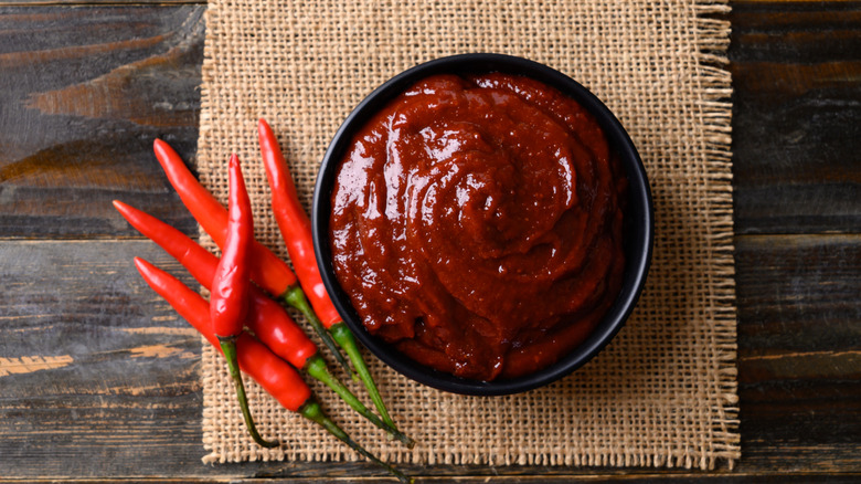 Gochujang paste in bowl on top of burlap beside chile peppers on a wooden table