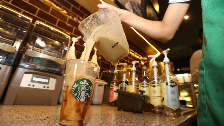 A Starbucks employee pours milk into a plastic Starbucks cup, with bottles and equipment in the background
