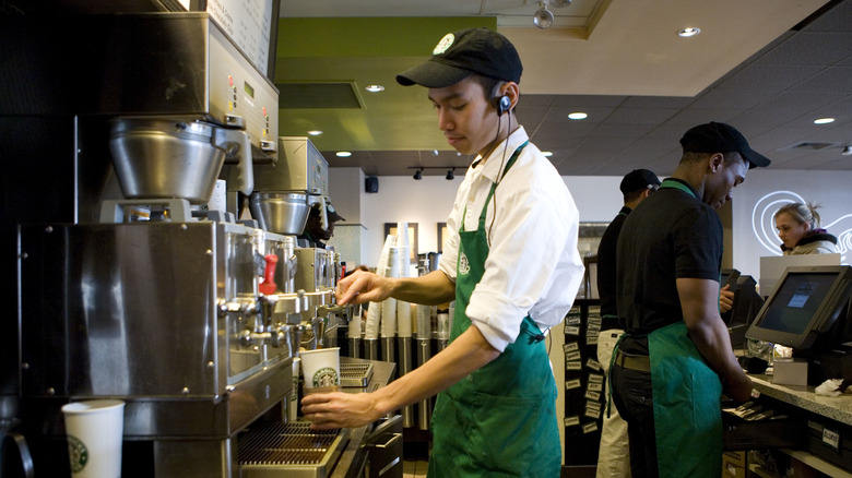A Starbucks barista prepares a beverage in a Starbucks branded cup, while other employees serve at the counter behind him