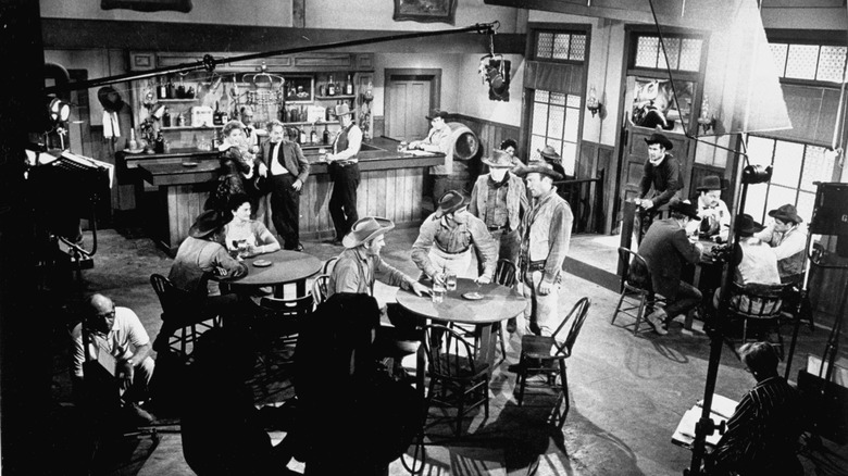 A black-and-white photograph of actors on the set of "Gunsmoke", with crew positioning lights, a boom microphone, and a camera in shadow.