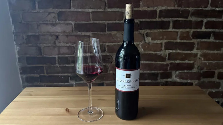 A glass of Charles Shaw merlot sits on a table next to an open bottle.