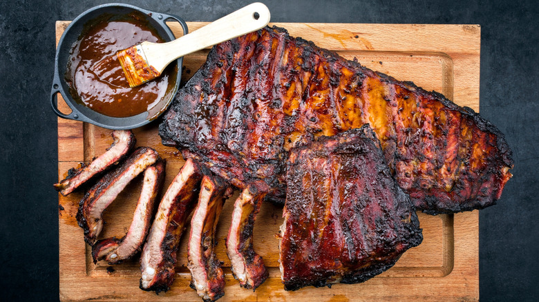 A slab of partially sliced St. Louis-style ribs sits on a wooden chopping board next to a bowl of barbecue sauce with a brush