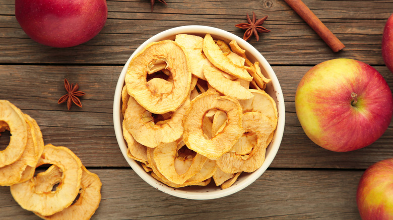 dried apples in a white bowl with star anise, cinnamon sticks, and more whole and dried apples on a wooden table.