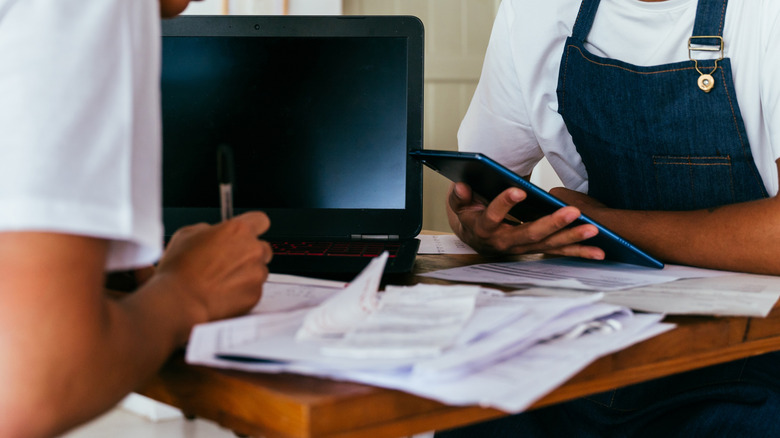 Two people wearing aprons work with paper, pen and computers