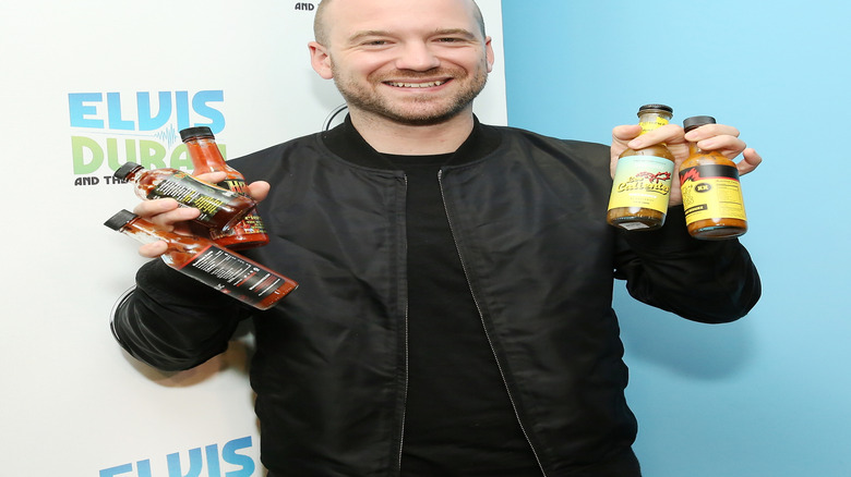 Sean Evins against Elvis Duran background holding hot sauce bottles in both sounds