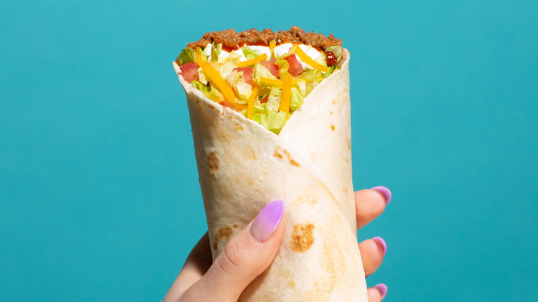 A hand with pale purple fingernails holding a burrito supreme against a turquoise background