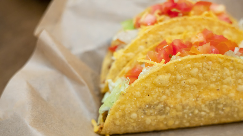 A pair of crunchy tacos topped with lettuce tomato and cheese sitting on a piece of paper