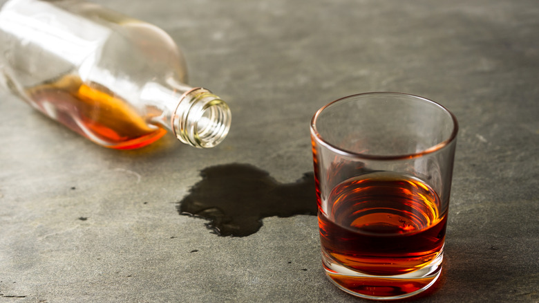 A glass of whiskey next to a spilled whiskey bottle on a grey surface