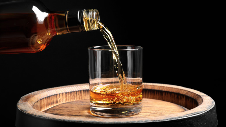 Bourbon whiskey being poured from glass bottle into rocks glass sitting on top of wood barrel