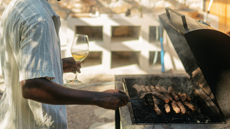A person grills sausages while holding a glass of white wine