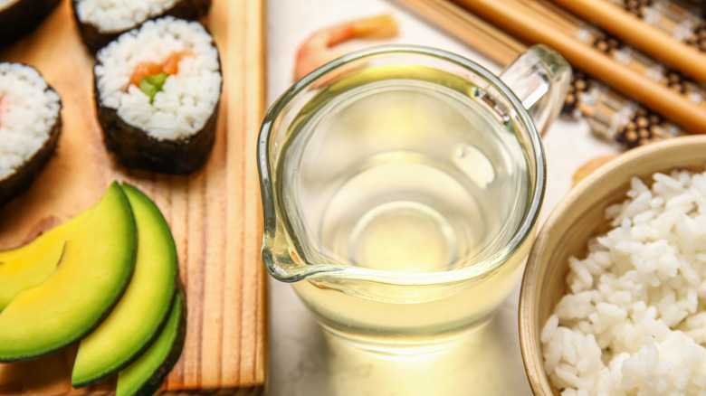 glass carafe of rice vinegar beside bowl of rice and cutting board with sushi and sliced avocado on it