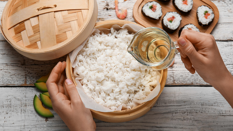 hands preparing to pour rice vinegar into basket of steamed rice