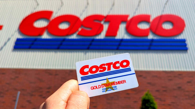 Hand holding Costco membership card in front of Costco sign on building