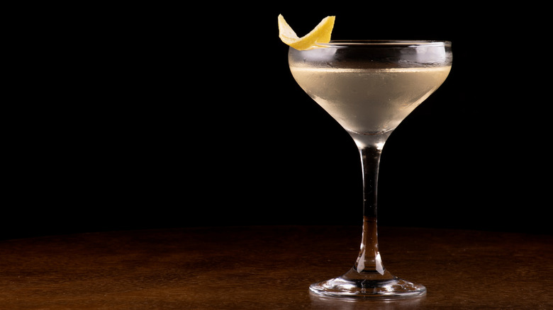 A coupe glass is filled with a slightly yellow cloudy liquid and topped with a lemon peel against a dark black and brown background