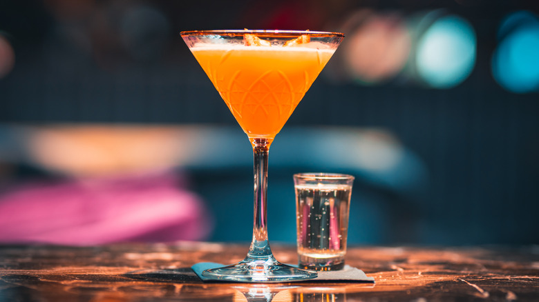 Jewel-toned fuzzy lights in shades of pink, orange, and blue glow in the background. In the front in clear focus is a single martini glass filled with a golden liquid and topped with a fruit. A single small shot of a bubbling liquid is next to the martini glass