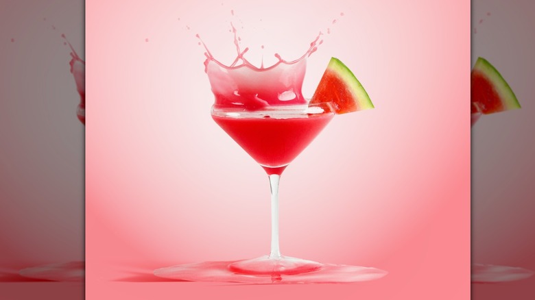 A watermelon wedge sits on the rim of a martini glass while a watermelon-red liquid is captured splashing out of the martini glass against a pink background