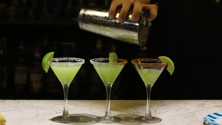 A hand holding a silver shaker is seen pouring a green liquid into three martini glasses with salt and tajin rims and lime wedges