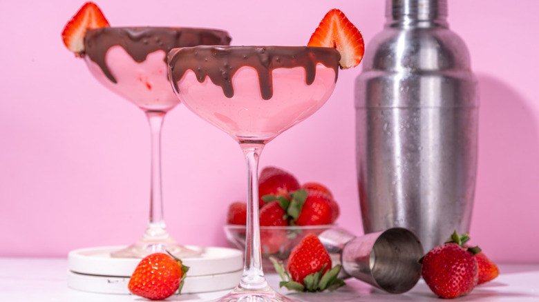 Fresh red strawberries stand out against a bright pink background. There are two martini glasses with chocolate dipped rims topped with a strawberry wedge. Fresh red strawberries are in a bowl and loose around the two martini glasses. A silver shot measurement tool and shaker are featured in the background