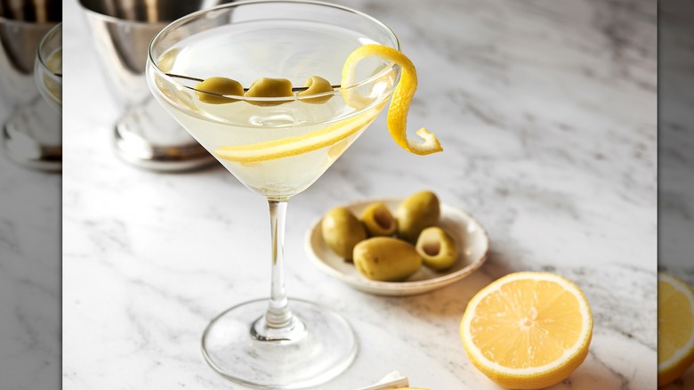 A silver shaker is seen in the background behind a single martini glass filled with a clear liquid topped with a lemon peel and olives. A small plate with additional green olives sits next to the martini glass and a half lemon wedge.