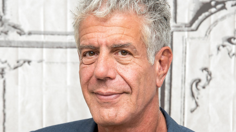 Anthony Bourdain in a gray jacket in front of a off-white and gray wall