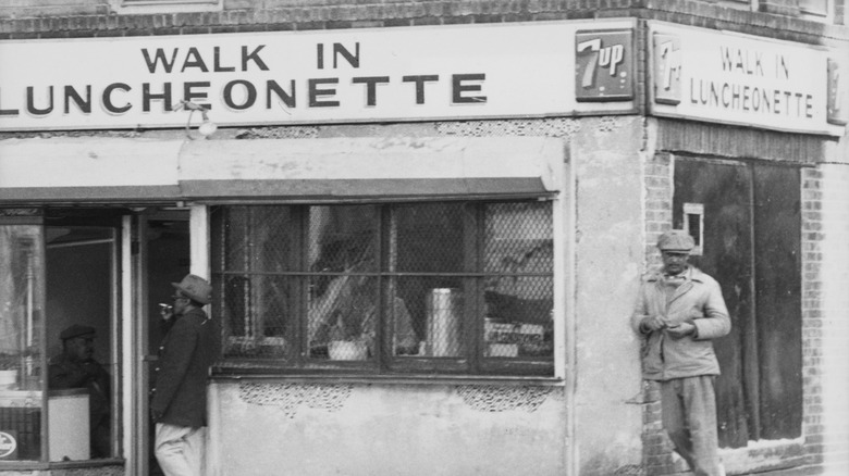 A black-and-white photograph of the exterior of a luncheonette, with a sign reading 'Walk In Luncheonette', with several patrons standing outside