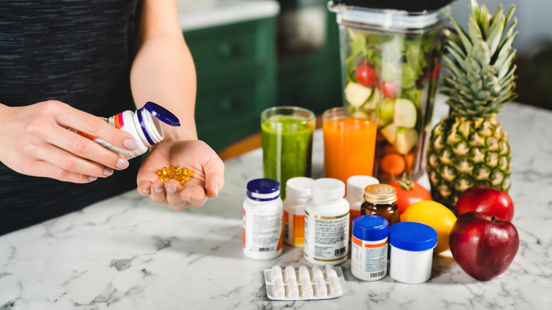 A person shakes out some vitamins from a bottle in front of an array of supplements, fruits, juices, and a blender full of vegetables