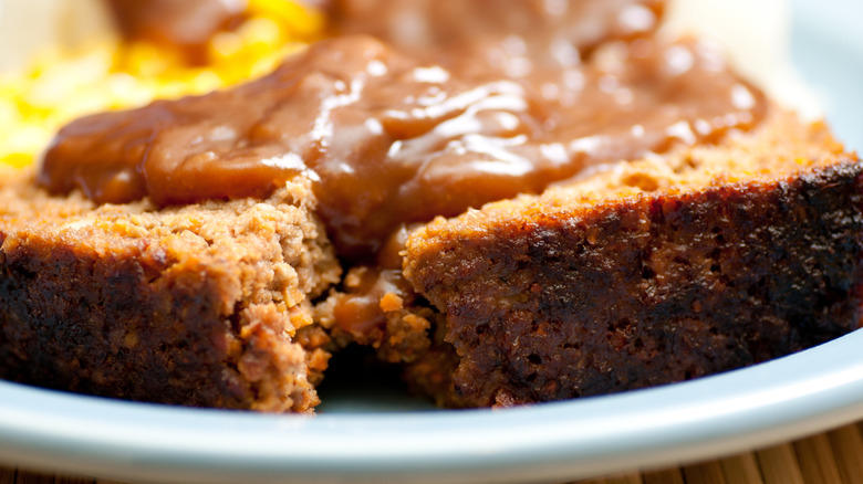a slice of meatloaf topped with gravy on a white plate with corn and mashec potatoes blurry in the background
