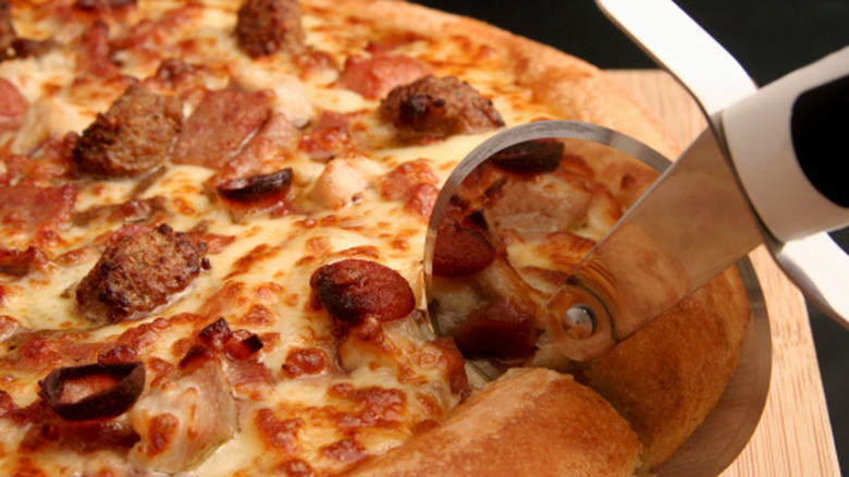 Close-up shot of a pizza cutter slicing through a pizza with pepperoni and sausage on it.