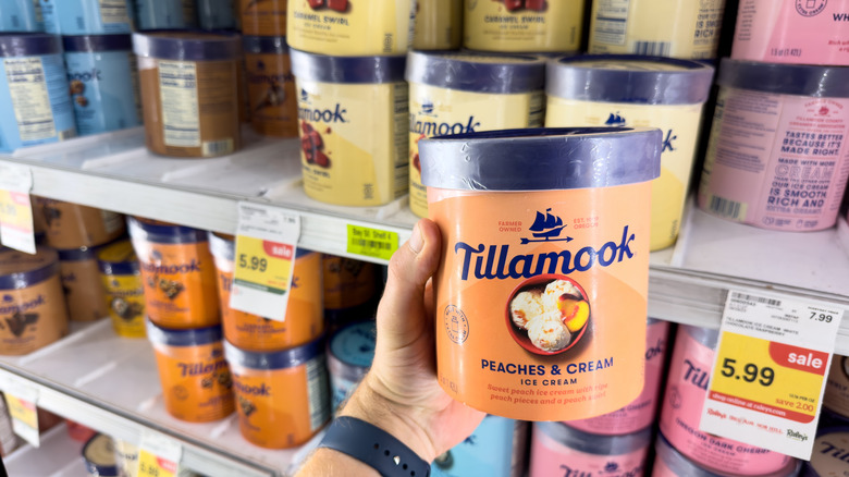 A close up of a person 's hand holding a pint of Tillamook Peaches & Cream in front of a freezer full of other TTillamook flavors.