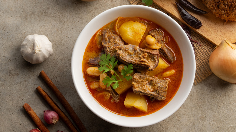 a bowl of curry in a red sauce with onions, garlic, chiles, and spices around it