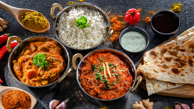 metal bowls of Indian curries and rice surrounded by spices, whole ingredients, and naan