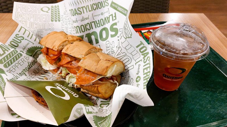 A Quiznos sub in a paper-lined basket next to a drink on a green tray
