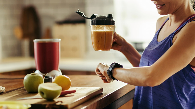 Qoman checking her watch and holding a smoothie standing next to cutting board with apples