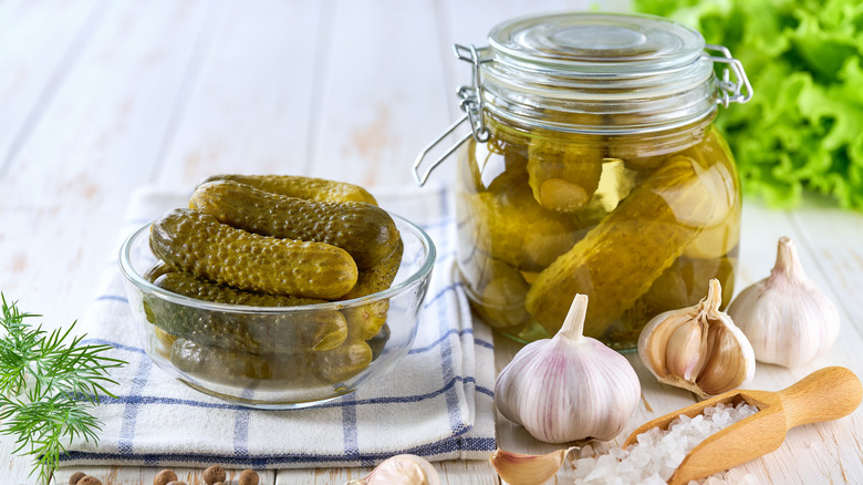 pickles in a bowl on a folded napkin next to a jar of pickles, heads of garlic, and large salt in a wooden spooon.