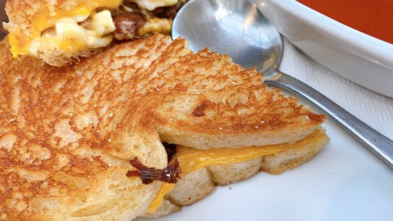 Grilled cheese filled with macaroni alongside spoon and bowl of tomato soup