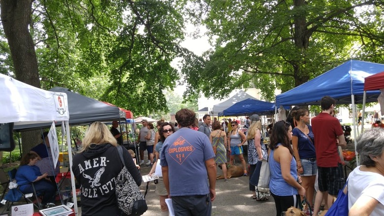Shoppers browsing farmers market tents underneath shade of trees