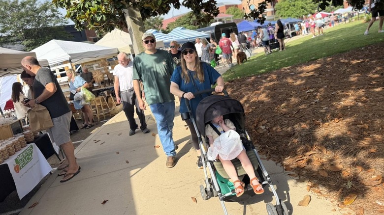 Woman pushing stroller along sidewalk beside farmers market tents