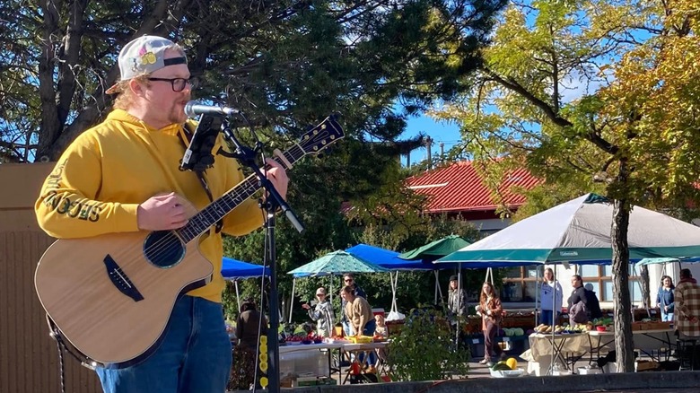 Man playing guitar while shoppers peruse farmers market tents in background
