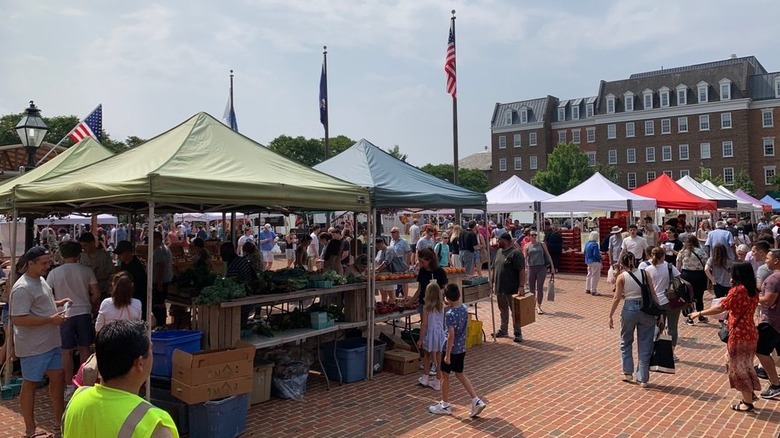 Shoppers exploring farmers market on brick expanse, flagpoles prominent in background