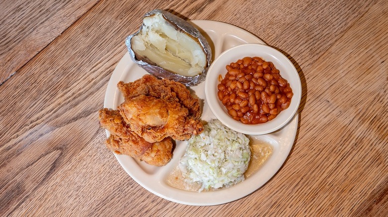 Top-down view of plate of fried chicken, coleslaw, baked potato, and baked beans on wooden table