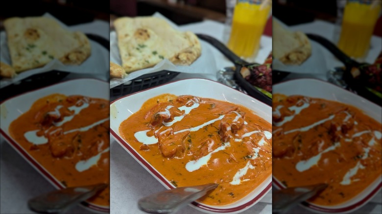 Butter chicken in serving dish with naan and mango lassi in background