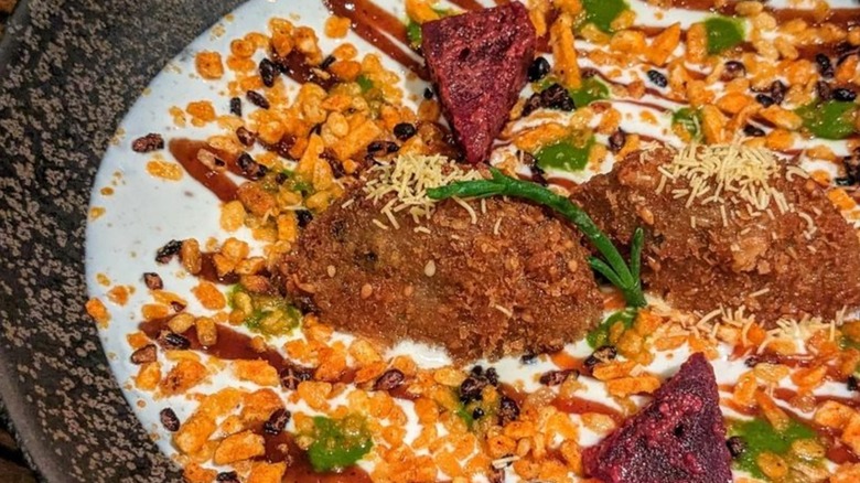 Two pieces of fried lamb in yogurt sauce, topped with crisps, chutneys, and two chunks of baked masala beet
