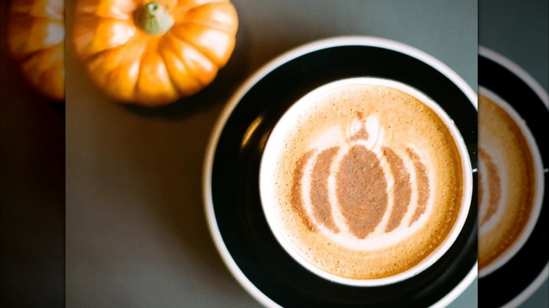 Pumpkin pie latte in mug, topped with pumpkin latte art, alongside decorative pumpkin on table