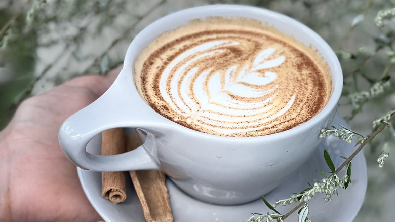 Hands holding saucer topped with mug of pumpkin pie latte, with two cinnamon sticks