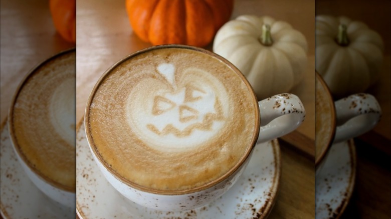 Pumpkin spice latte with pumpkin latte art in mug, alongside two pumpkins on table
