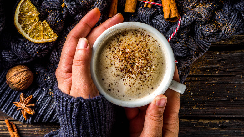 Hands cradling pumpkin spice latte amidst spices and blanket