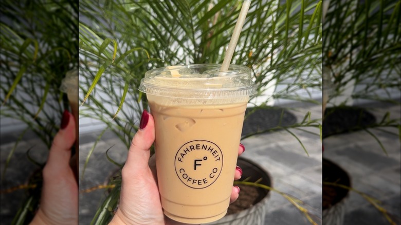 Hand holding pumpkin spice latte in plastic to-go cup in front of plant